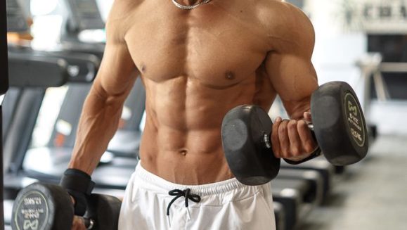 A shirtless, muscular man exercises with dumbbells in a well-lit gym, focusing on bicep curls.