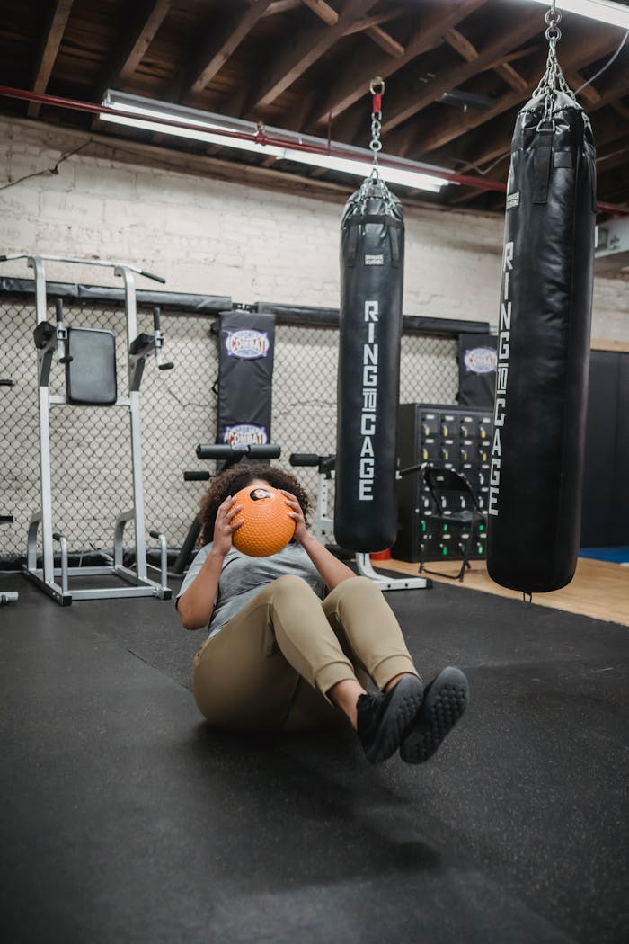 Full body of unrecognizable plus size ethnic female with Afro hair in sportswear lying on floor while doing abs exercise with medicine ball in modern gym